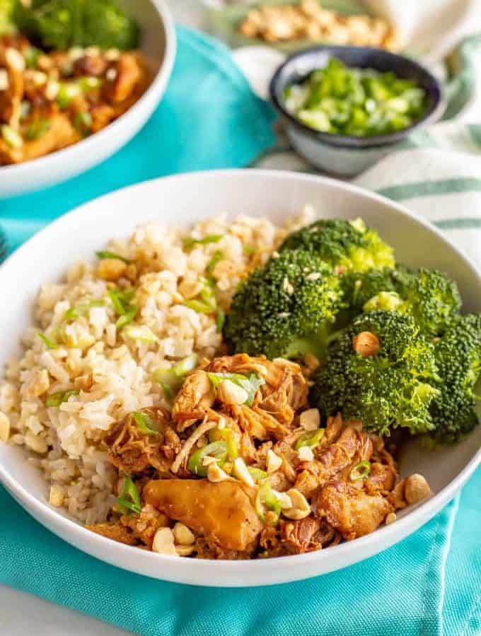 A white bowl with brown rice, honey garlic chicken and steamed broccoli arranged with green onions and peanuts sprinkled on top.