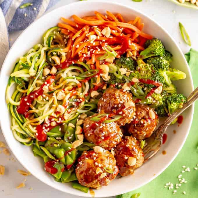 A white bowl full of spiralized veggies, broccoli and snow peas and Asian turkey meatballs with a serving fork resting to the side.