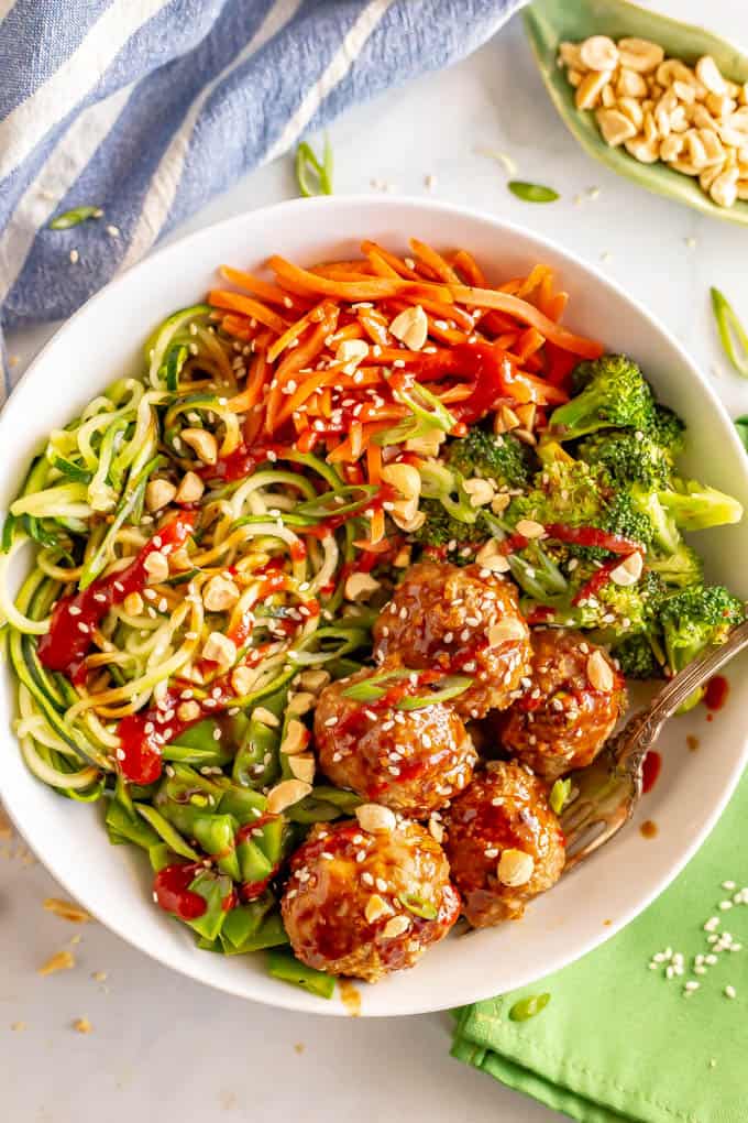 A white bowl full of spiralized veggies, broccoli and snow peas, Asian turkey meatballs and crushed peanuts sprinkled on top with a serving fork resting to the side.