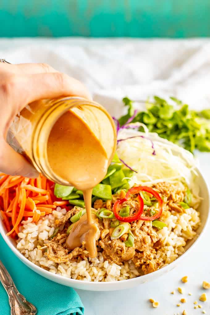 A hand pouring a homemade peanut dressing over a grain bowl of rice, peanut chicken and fresh veggies.