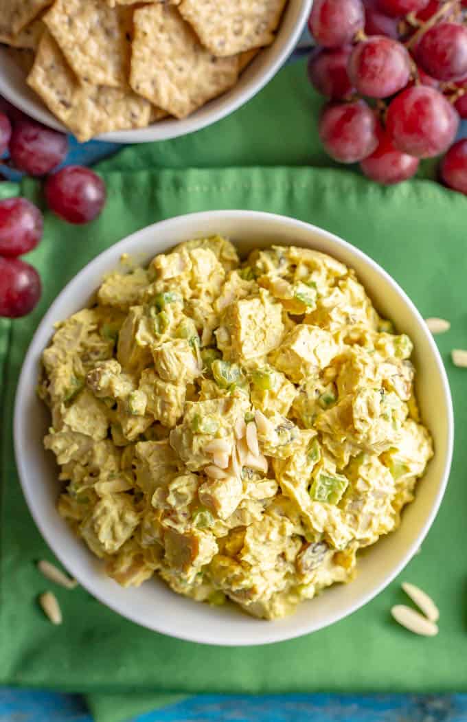 Healthy curried chicken salad in a white bowl with red grapes and crackers nearby