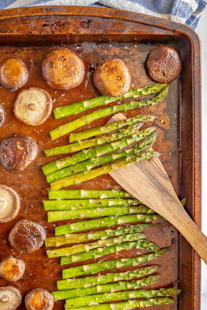 A wooden spatula resting on a dark sheet pan of roasted asparagus and mushrooms.