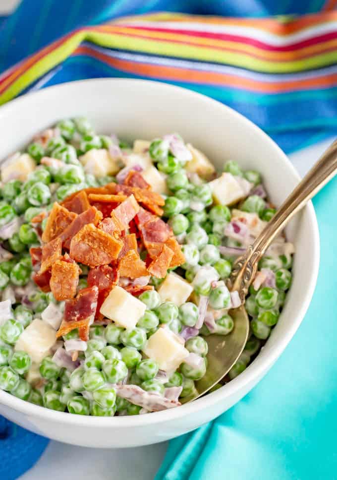 A spoon resting in a small white bowl full of a creamy pea salad with red onion, small squares of cheese and cooked, crumbled bacon on top.