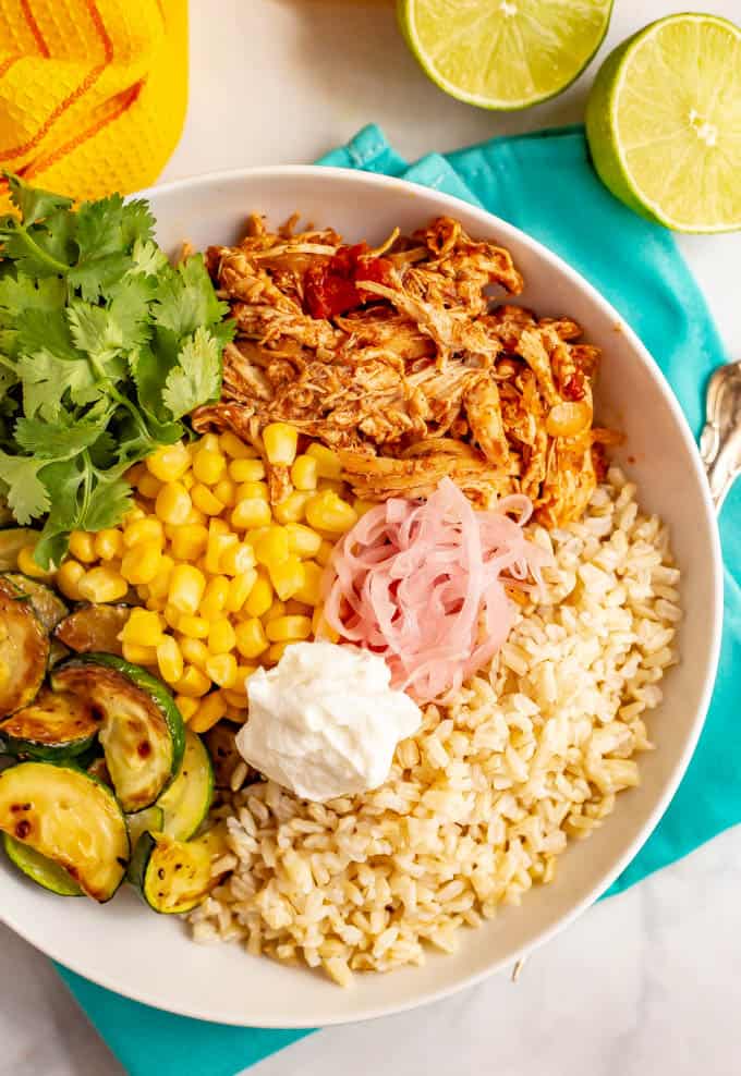 A bowl with piles of rice, shredded salsa chicken, corn, charred zucchini and garnishes of cilantro, pickled red onions and sour cream.