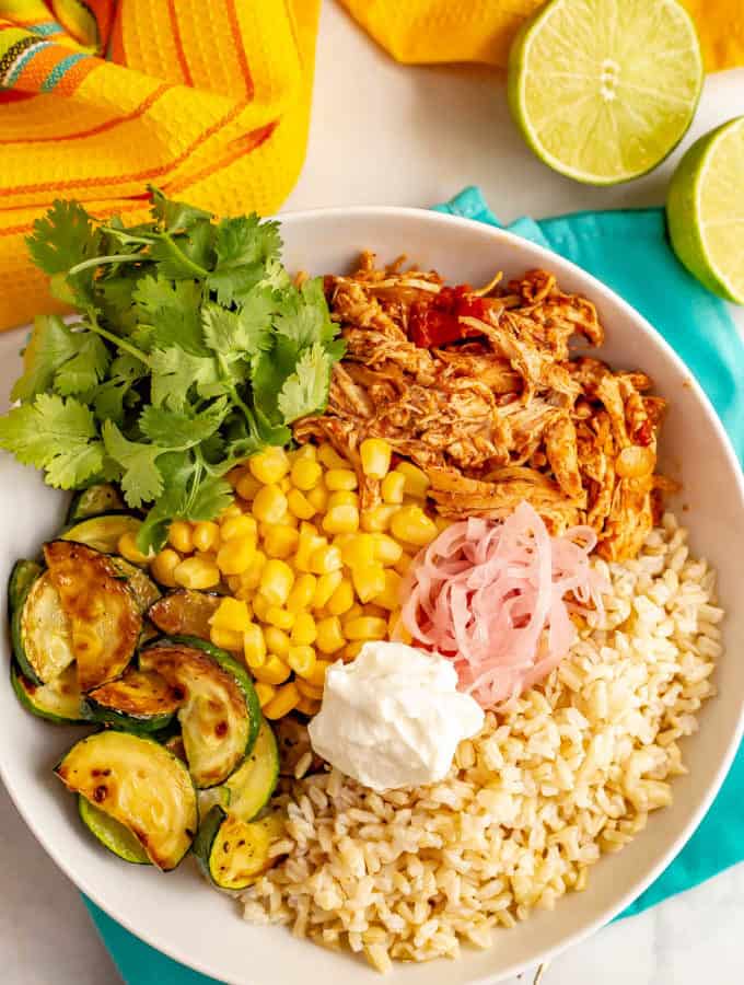 A grain bowl arranged with steamed brown rice, shredded salsa chicken, corn, roasted zucchini with pickled red onions and sour cream dolloped on top and some cilantro sprigs to the side.
