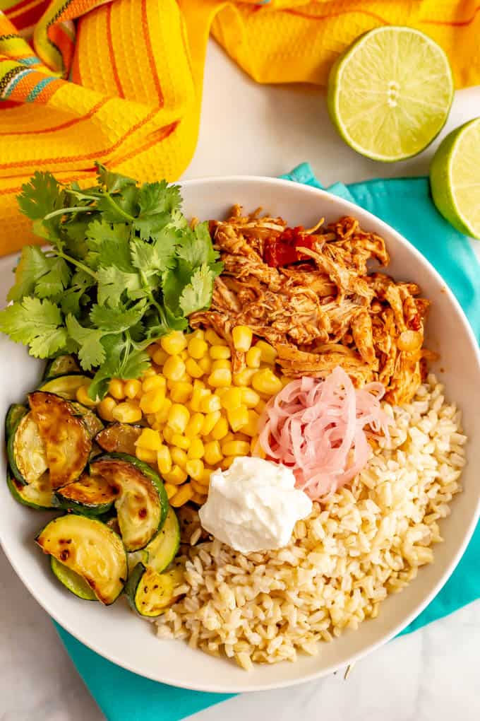 A grain bowl arranged with steamed brown rice, shredded salsa chicken, corn, roasted zucchini with pickled red onions and sour cream dolloped on top and some cilantro sprigs to the side.
