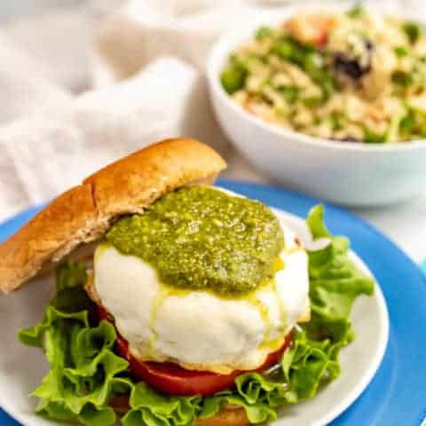 A cheese smothered burger on a bun with pesto, lettuce and tomato and a salad in a bowl in the background