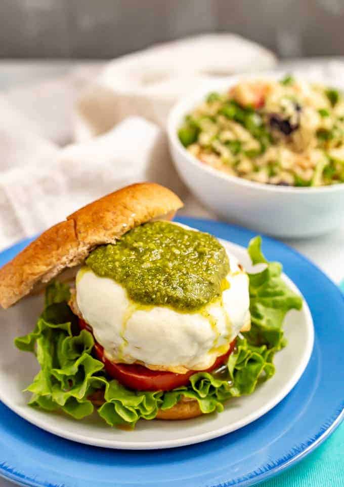 A cheese smothered burger on a bun with pesto, lettuce and tomato and a salad in a bowl in the background