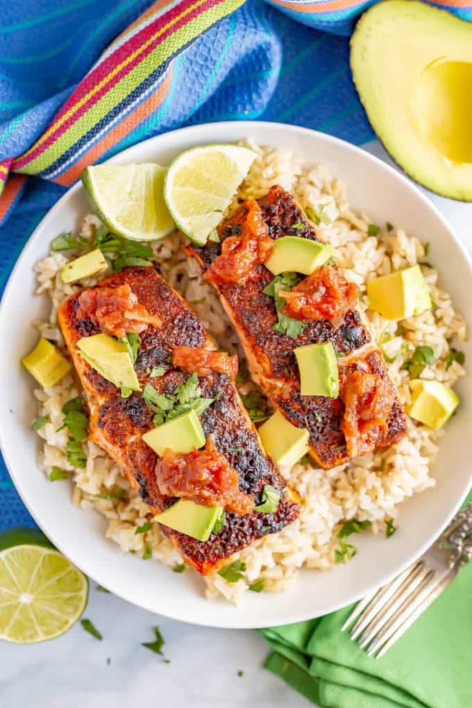 A bowl of rice topped with two filets of southwest baked salmon topped with salsa and diced avocado with lime wedges to the side.