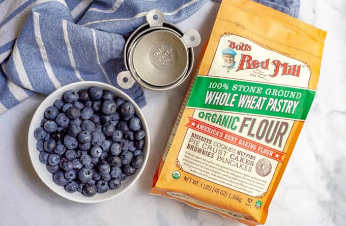 Whole wheat pastry flour, blueberries and baking measuring cups laid out with a blue throw towel