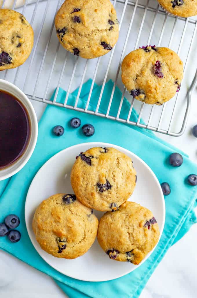 Overhead of fluffy blueberry muffins on a white plate with extras on a wire rack and blueberries scattered around