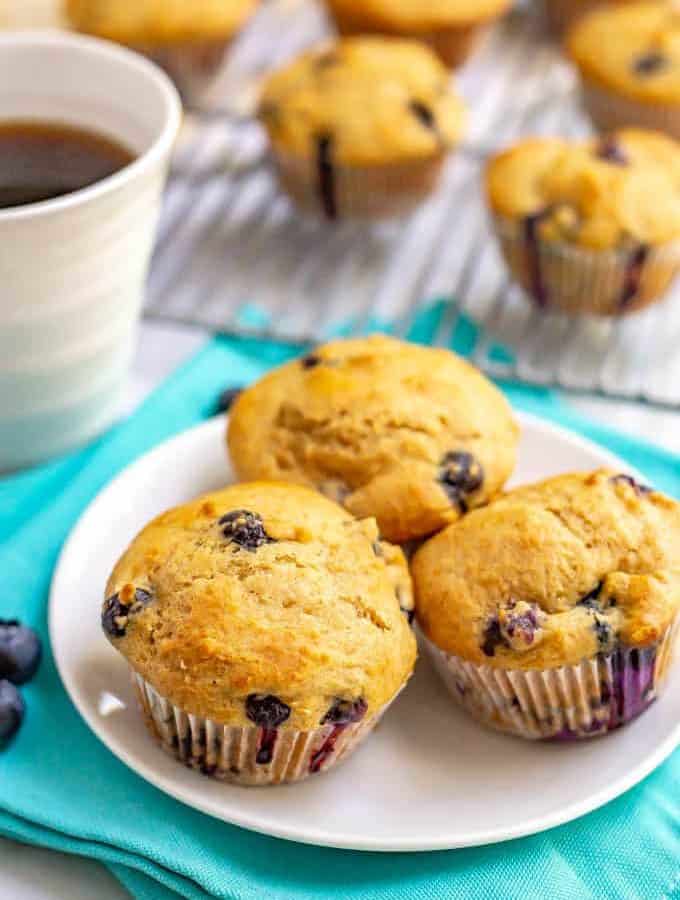 A trio of homemade healthy blueberry muffins on a small white plate with a cup of coffee and more muffins on a cooling rack in the background.