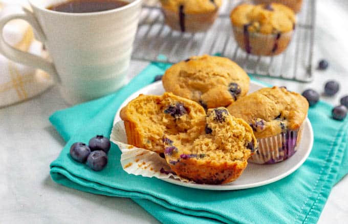 Blueberry muffins on a white plate with coffee and extra muffins on a muffin rack