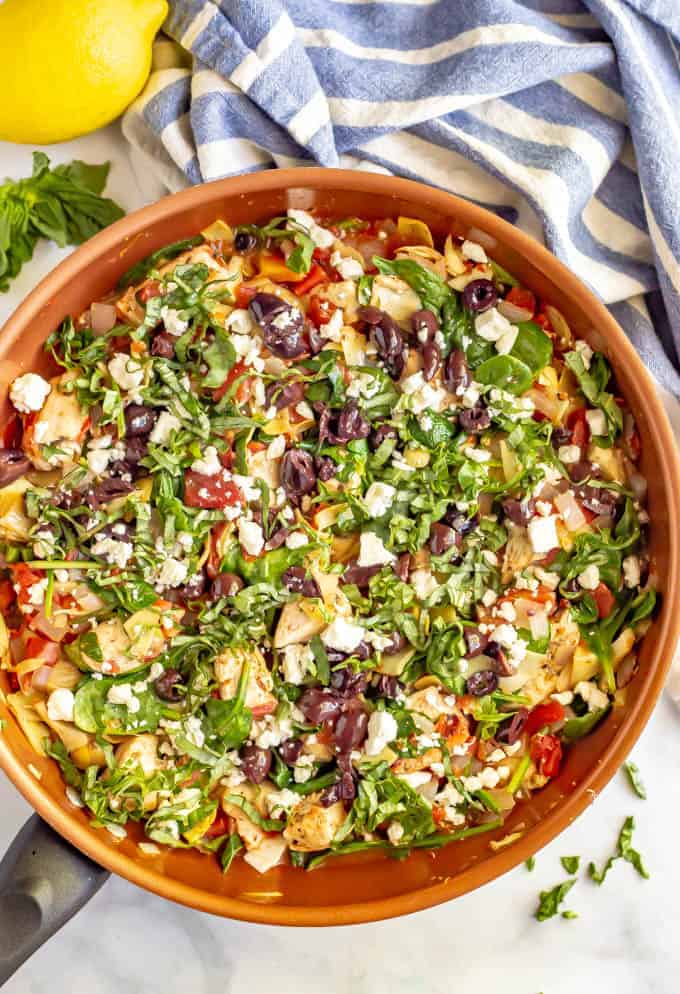 Full copper skillet of chicken and vegetables with basil on top and nearby and a lemon beside the pan