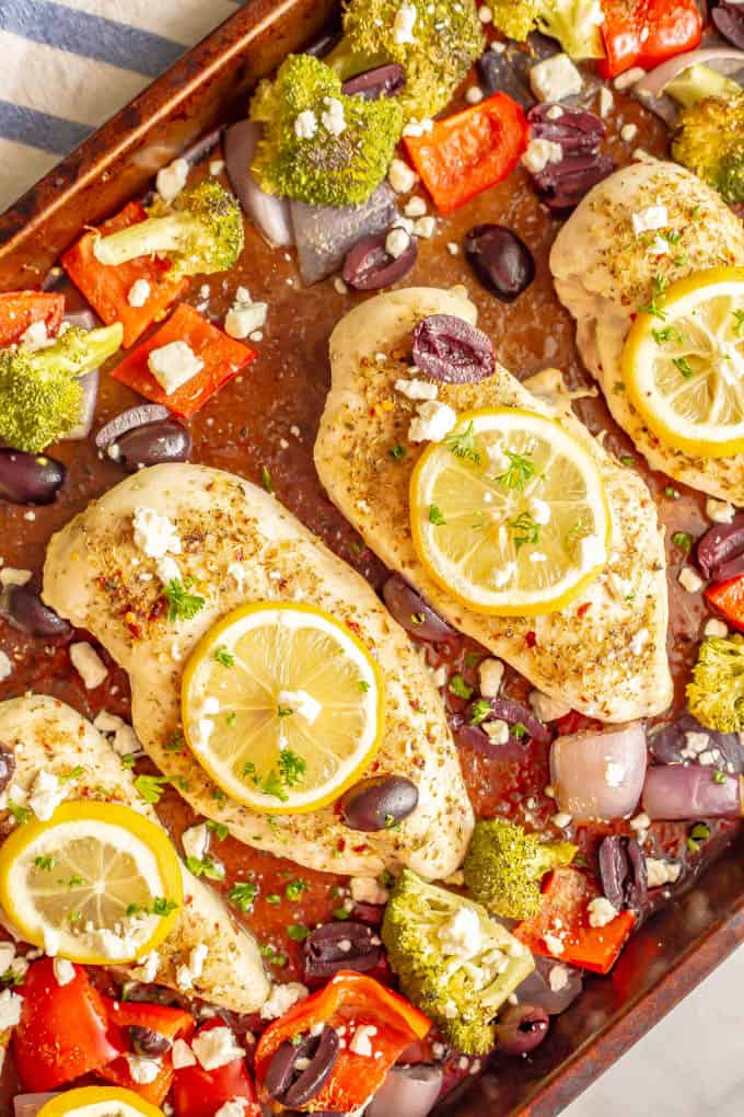 Chicken breasts on a sheet pan with lemon slices on top and broccoli, peppers, red onions, olives and feta cheese scattered around.