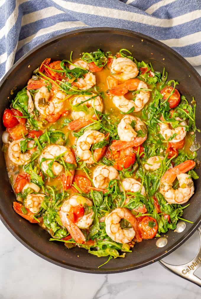 Shrimp, arugula and tomatoes cooking in a large pan with a blue striped kitchen towel nearby