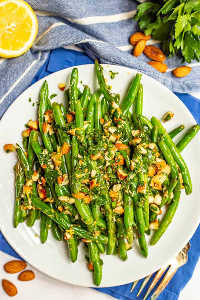 Green beans served on a plate with a sprinkling of almonds mixed with parsley and lemon juice and garnishes nearby