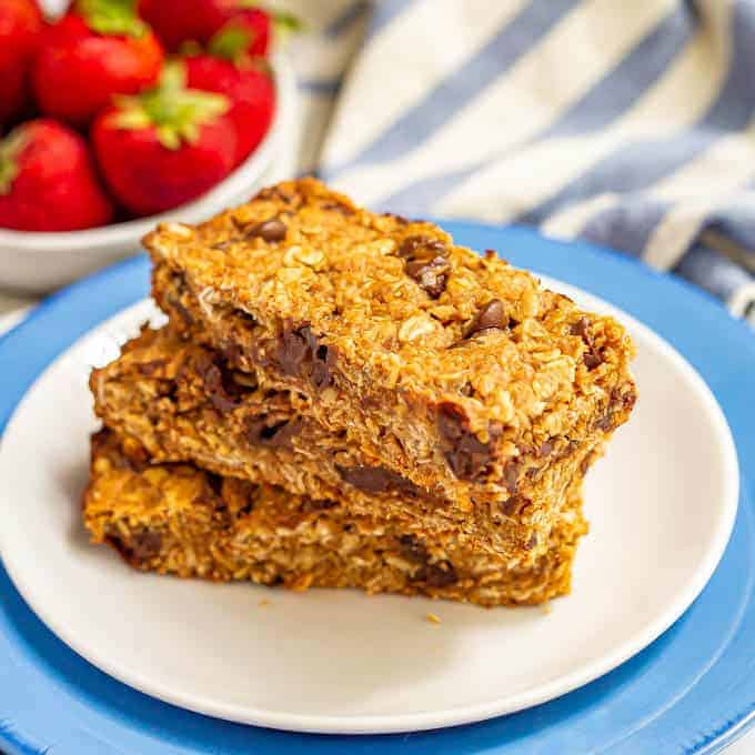 Three thick, chewy homemade granola bars stacked on a white plate with strawberries in the background
