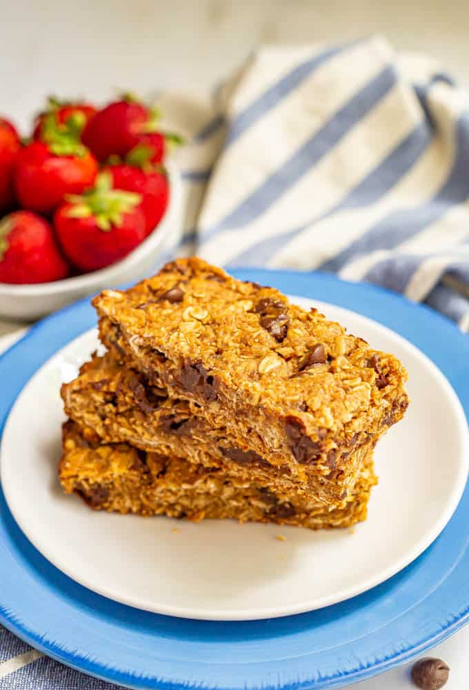 Three chocolate chip granola bars stacked on a plate with a bowl of strawberries in the background