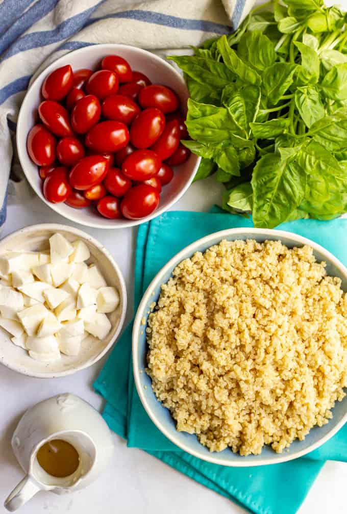 Separate bowls of tomatoes, cooked quinoa and mozzarella with a bunch of fresh basil and lemon vinaigrette nearby