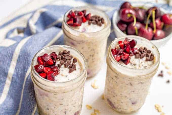 Cherry overnight oats in jars with mini chocolate chips on top and a bowl of cherries nearby