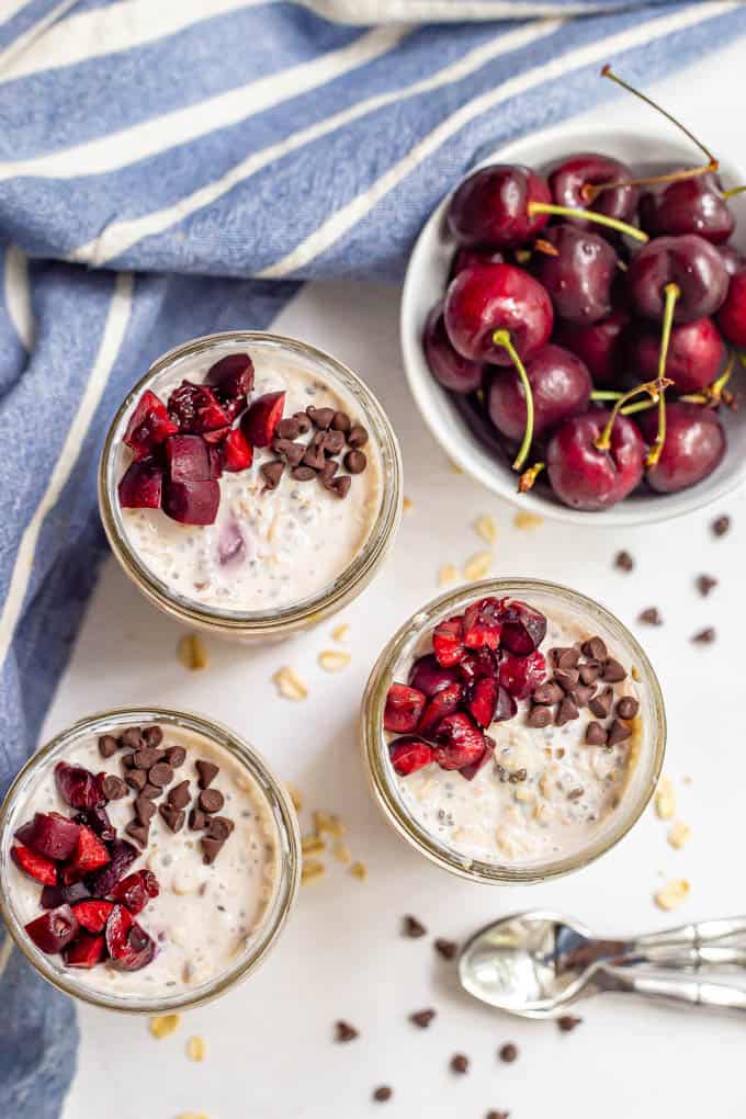 Overnight oats with fresh cherries and mini chocolate chips in small jars and a bowl of cherries nearby