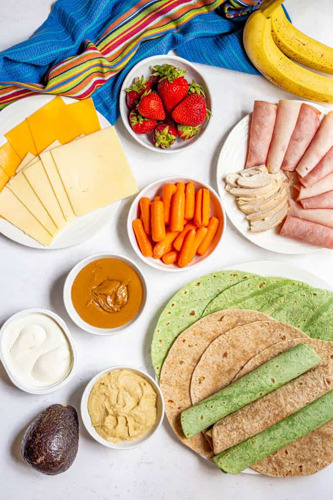 Ingredients laid out for making healthy lunch wraps, including deli meats, sliced cheese, peanut butter, cream cheese, hummus, avocado, banana and an assortment of wraps.