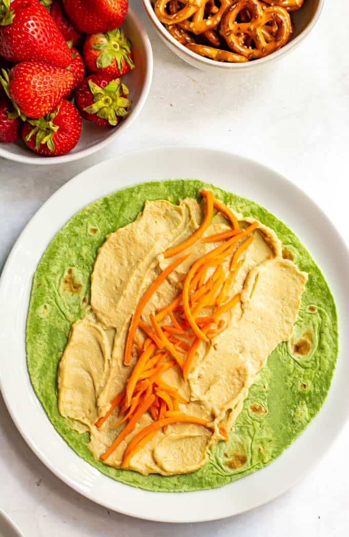 Hummus and shredded carrots on a spinach wrap with bowls of strawberries and pretzels in the background