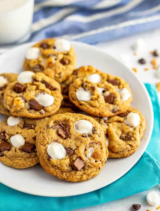 A white plate full of chocolate chip cookies with marshmallows and a glass of milk in the background