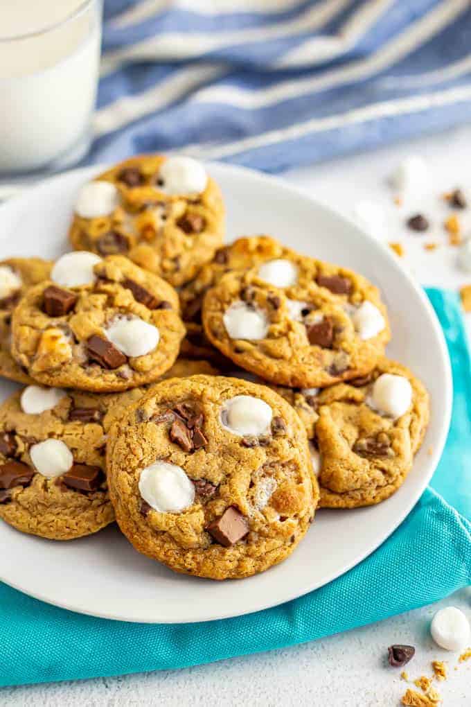 A white plate full of chocolate chip cookies with marshmallows and a glass of milk in the background