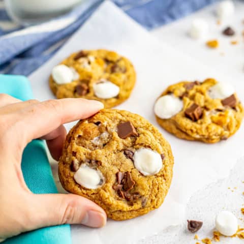 A hand holding a cookie with mini marshmallows and chocolate chunks on top