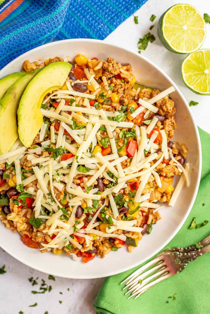 Bowl of ground turkey, rice, beans and veggies with cheese, cilantro and avocado served with two forks and limes nearby