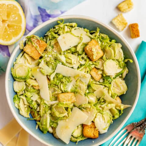 A serving bowl filled with Brussel sprout Caesar salad with a lemon, Parmesan cheese and croutons scattered nearby