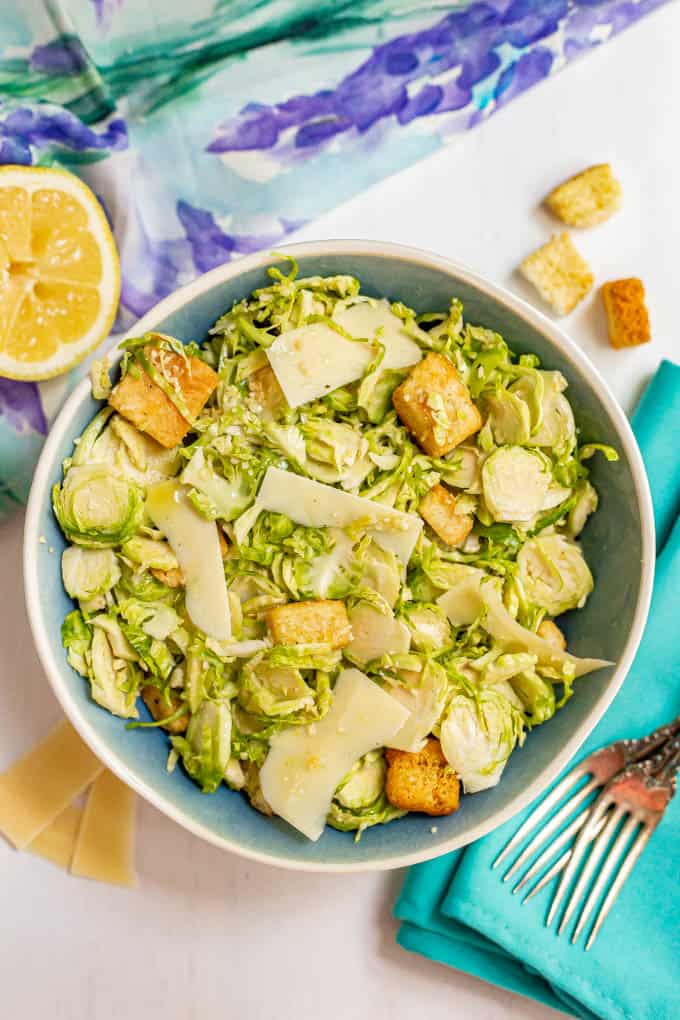 A serving bowl filled with Brussel sprout Caesar salad with a lemon, Parmesan cheese and croutons scattered nearby