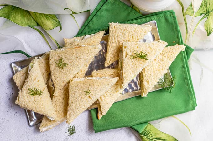 Overhead shot of classic cucumber sandwiches stacked on a silver serving tray with garnishes of fresh dill