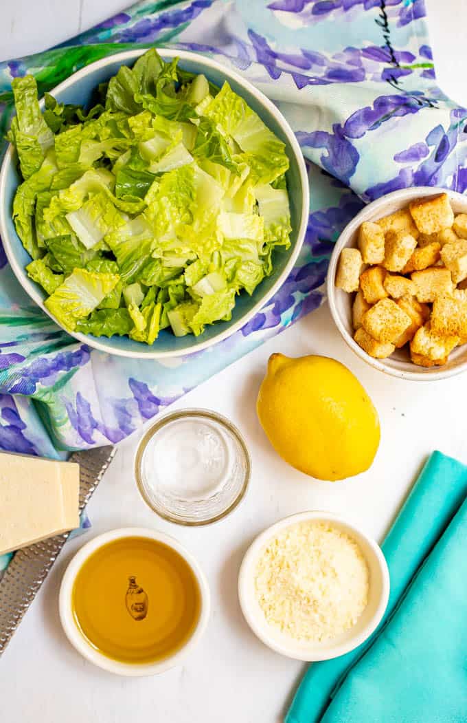 Ingredients for homemade Caesar salad dressing in separate containers with a large salad bowl of romaine in the background