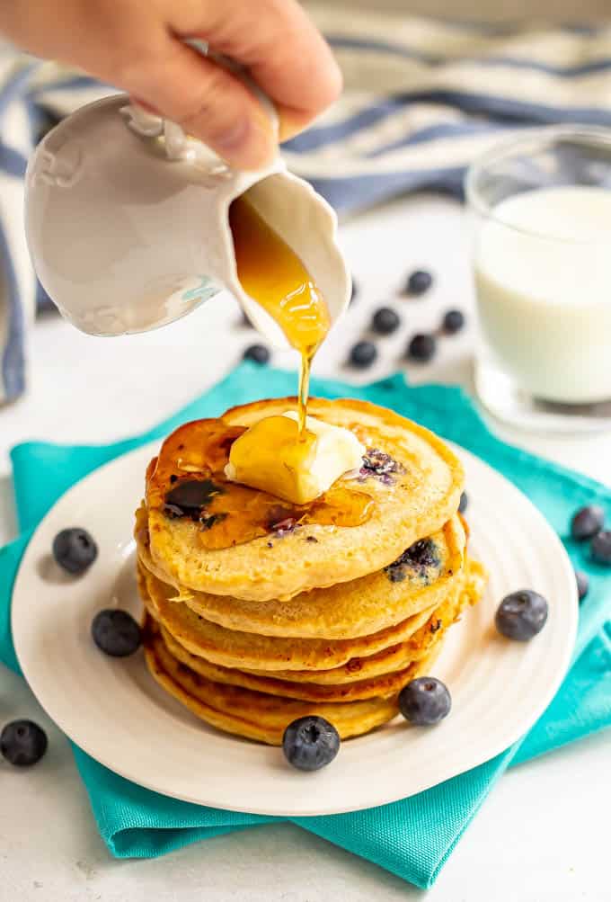 Maple syrup being poured over a stack of blueberry pancakes with blueberries scattered nearby