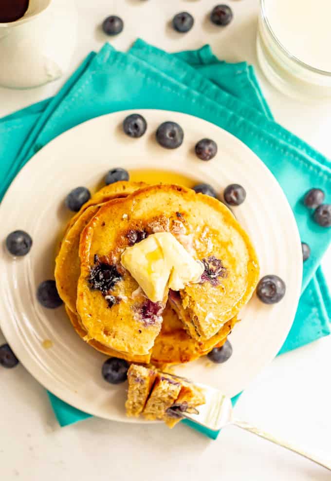 A top down shot of a stack of blueberry pancakes with butter and maple syrup and a forkful bite laying to the side