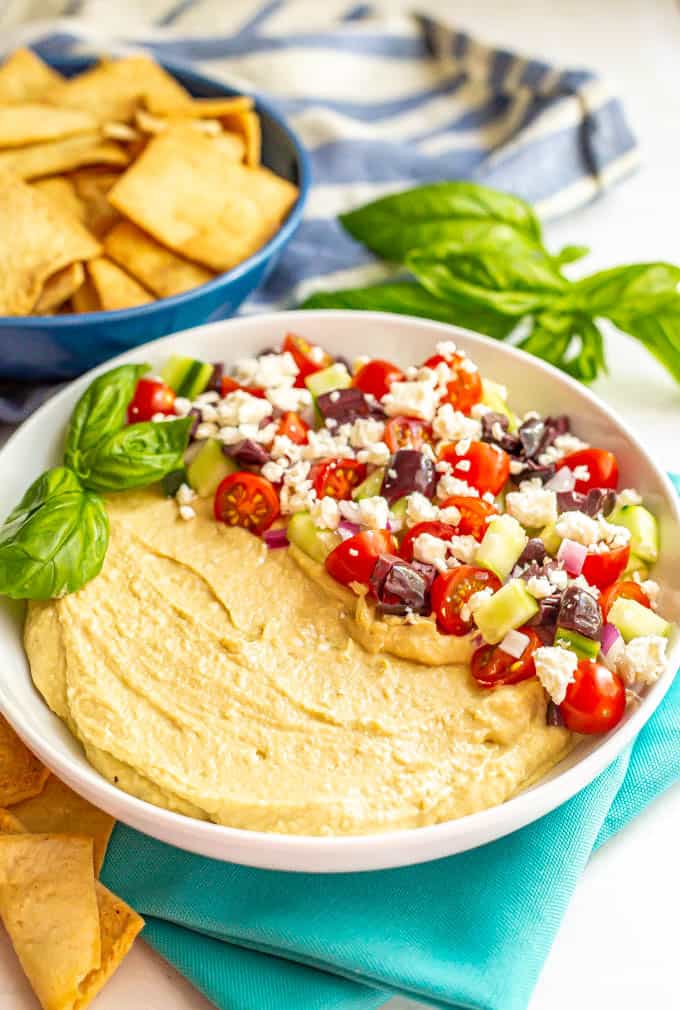 Layered hummus dip with tomatoes, cucumber, olives and feta cheese and a bowl of pita chips in the background