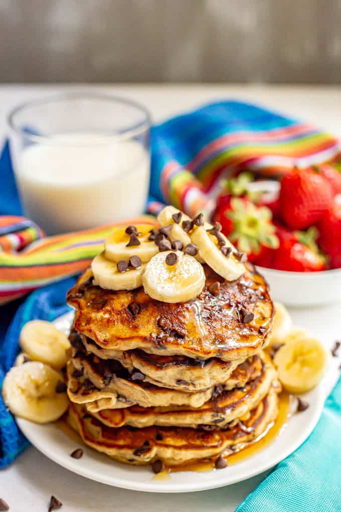 A stack of fluffy banana chocolate chip pancakes served on a white plate with sliced bananas, mini chocolate chips and maple syrup