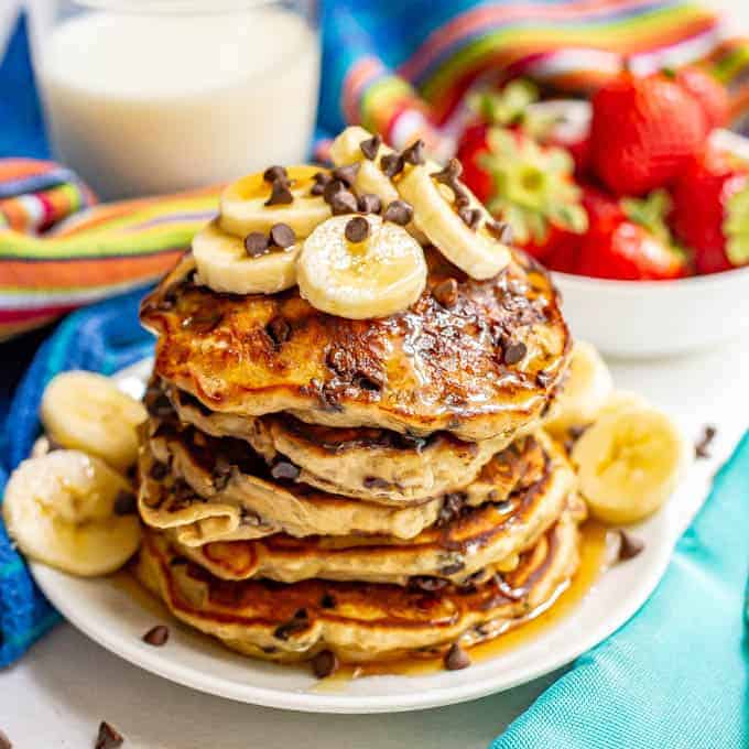 A stack of fluffy banana chocolate chip pancakes served with sliced bananas, mini chocolate chips and maple syrup