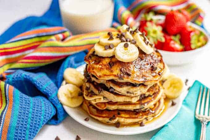 A stack of fluffy pancakes with chocolate chips served on a white plate with bananas and maple syrup