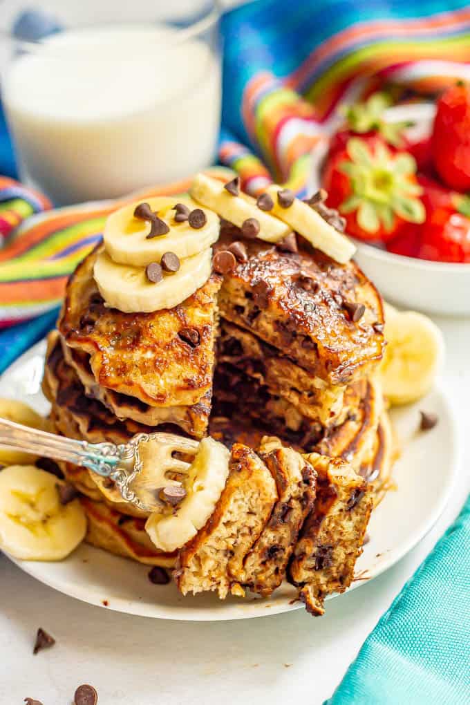 A forkful of pancakes with chocolate chips and banana being taken from a stack on a white plate