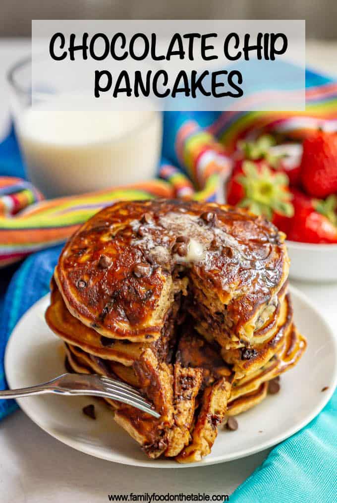 A fork piercing a bite of a stack of chocolate chip pancakes on a white plate with text on top of the photo.