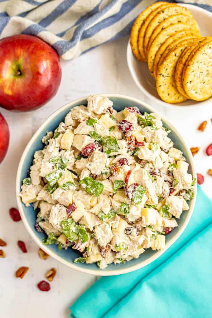 A serving bowl full of harvest chicken salad with apples, pecans, dried cranberries and spinach and crackers and apples nearby