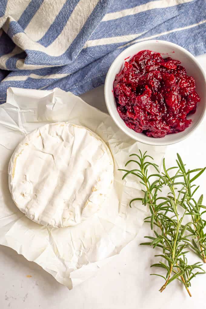 Brie cheese, cranberry sauce and fresh rosemary laid out on a table with a blue striped towel