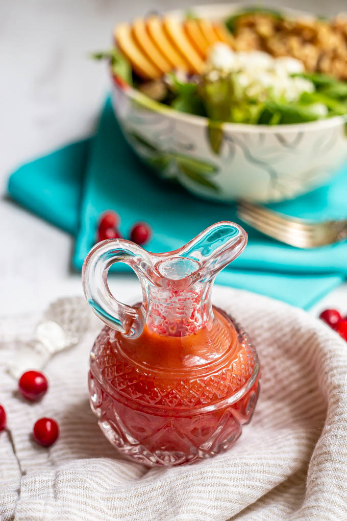 Bright cranberry dressing in a clear pouring glass with a salad bowl in the background