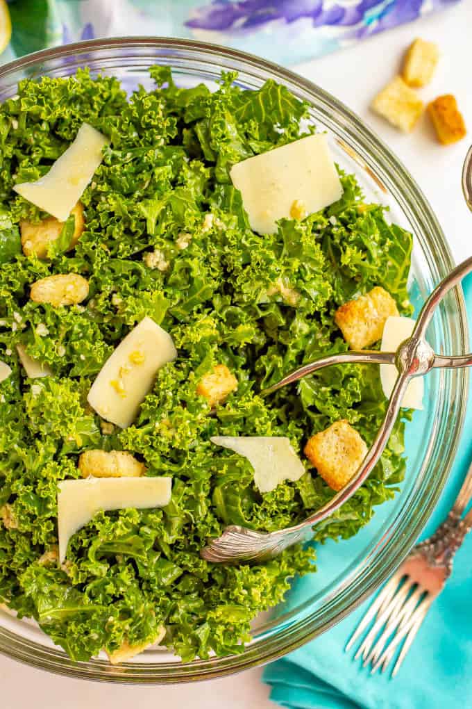 Silver salad tongs taking a scoop of kale Caesar salad from a large glass bowl