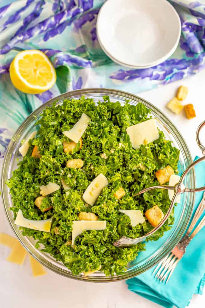 Silver salad tongs taking a scoop of kale Caesar salad from a large glass bowl with croutons and Parmesan cheese on the table