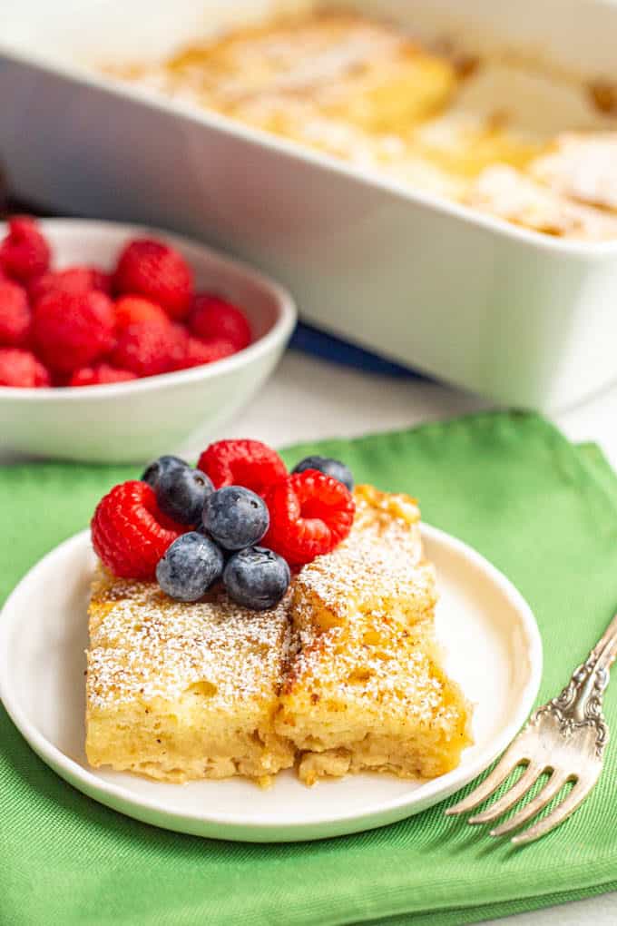 A slice of overnight French toast served on a white plate with fresh berries and a fork alongside